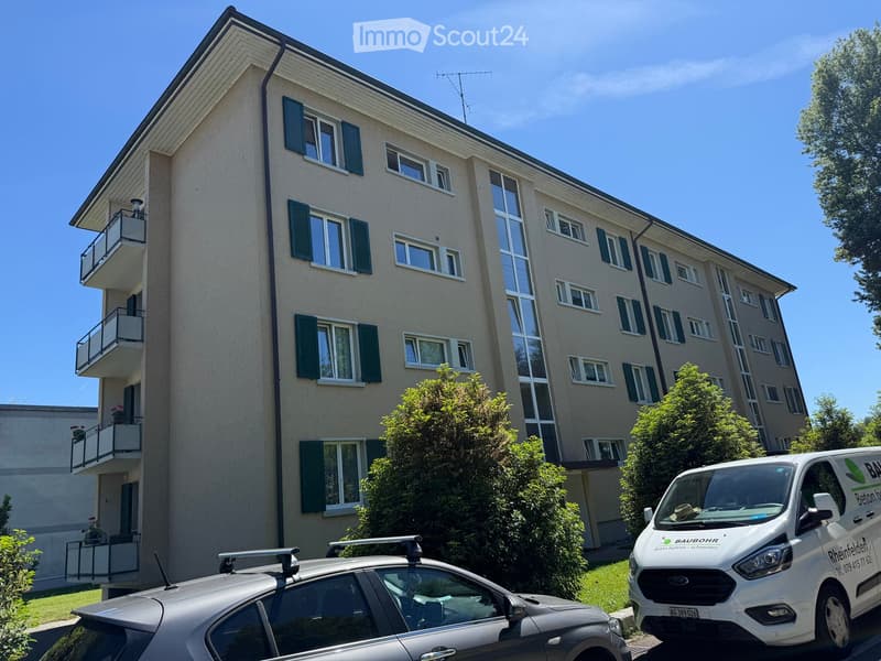 Four-story residential building with multiple windows, green shutters, and balconies. Two cars are parked in front, and there is a van with a logo on the side.