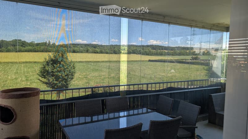Balcony with a view of a green field and trees, featuring a table and chairs.