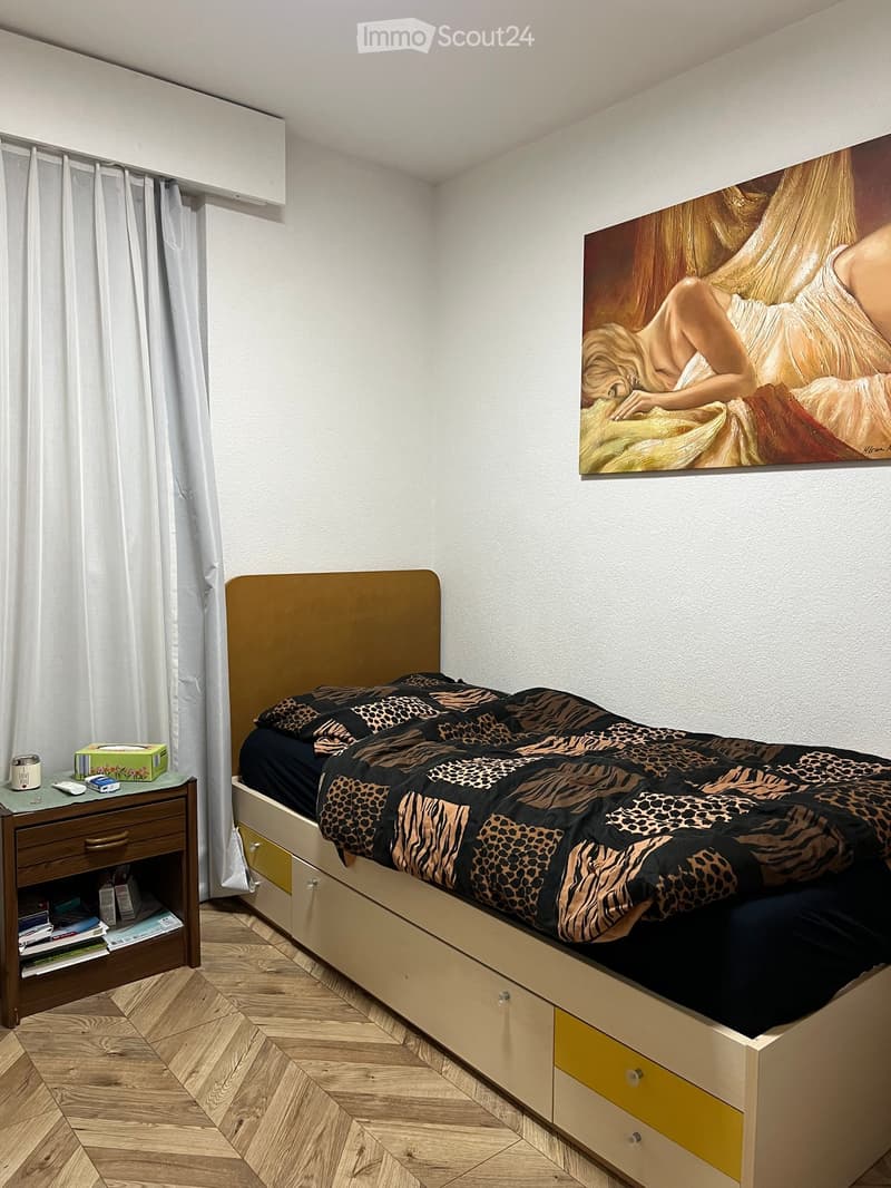 A modern bedroom with a large bed featuring a leopard print duvet, a wooden nightstand with books and a cup, and a large painting on the wall.