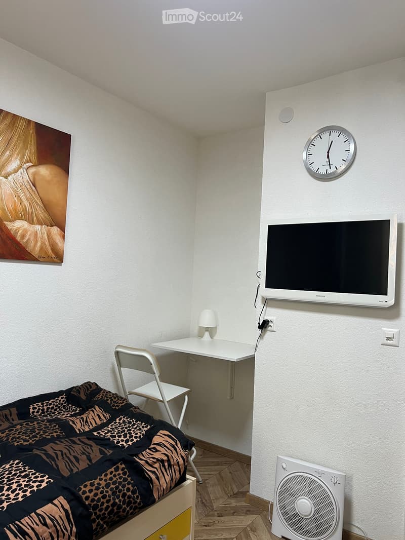 Cozy bedroom with a bed, desk, chair, lamp, fan, TV, and clock on the wall.