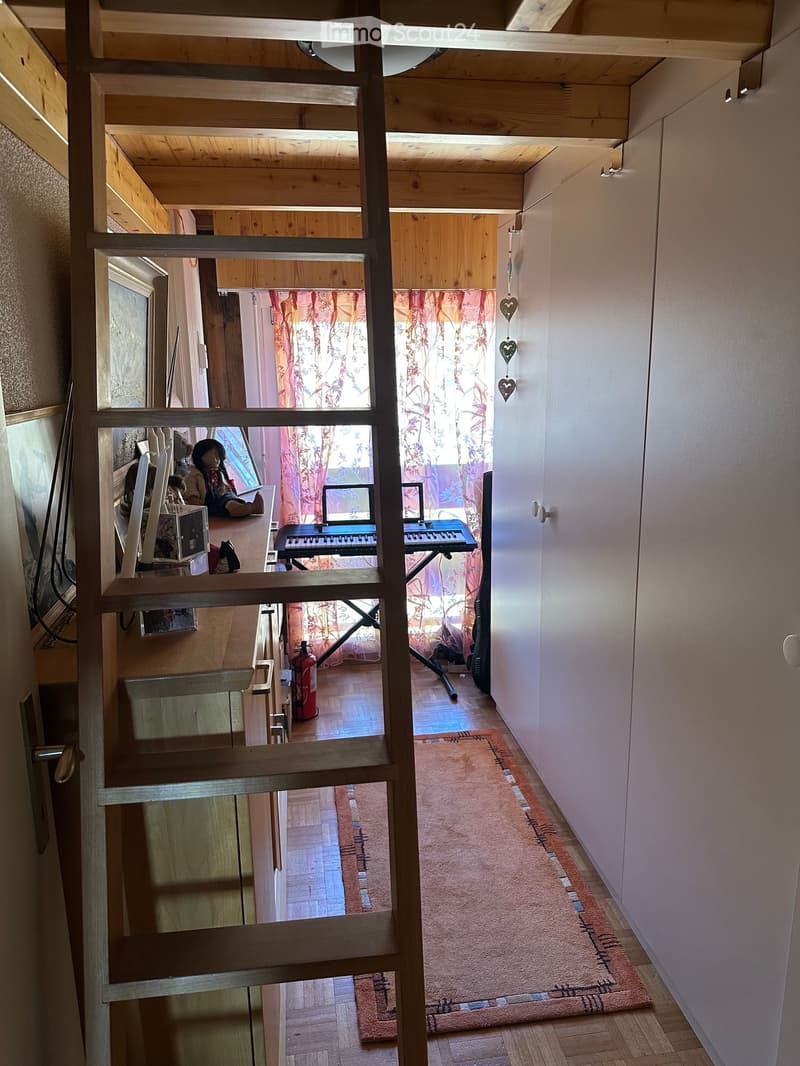 A narrow hallway with wooden beams and exposed ceiling beams, leading to a room with a keyboard, computer, and doll on a desk, and a fire extinguisher on the floor.