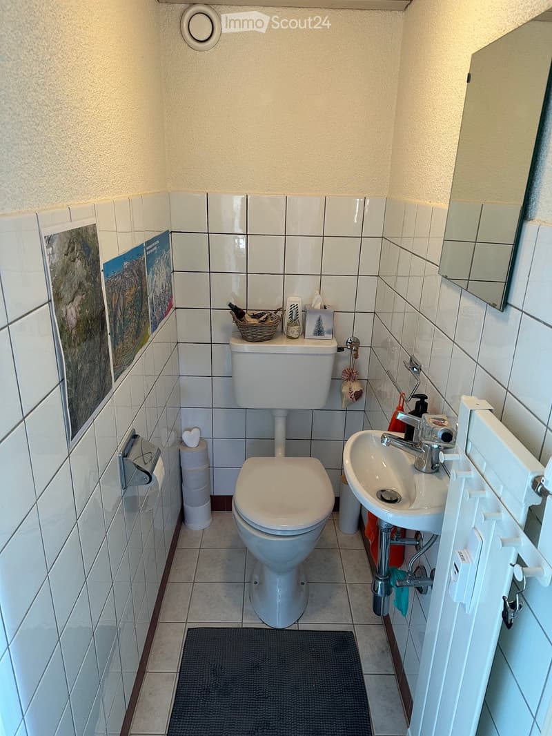 Compact bathroom with white tiled walls and floor, featuring a toilet, sink with chrome faucet, mirror, toilet paper holder, and a small basket on the toilet tank.