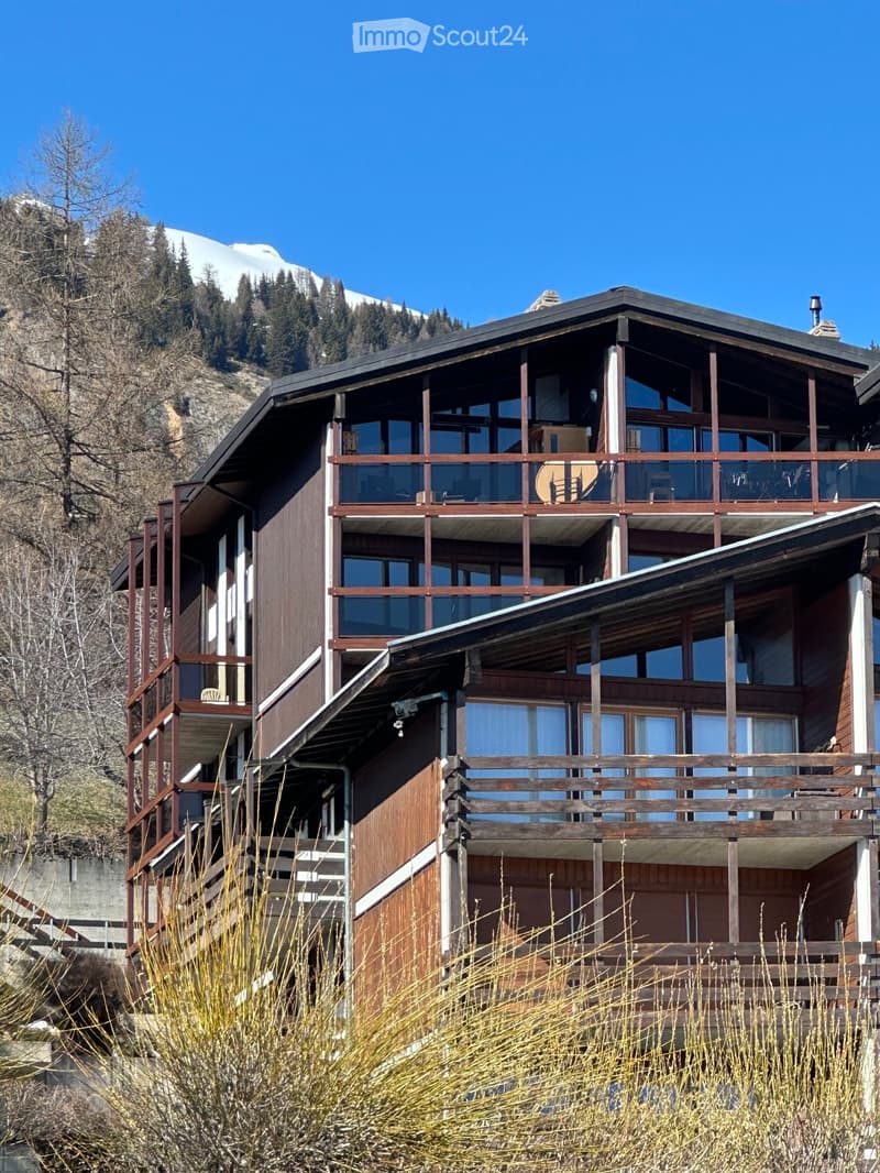 A two-story wooden house with large windows and balconies, situated in a mountainous area with snow-capped peaks in the background.