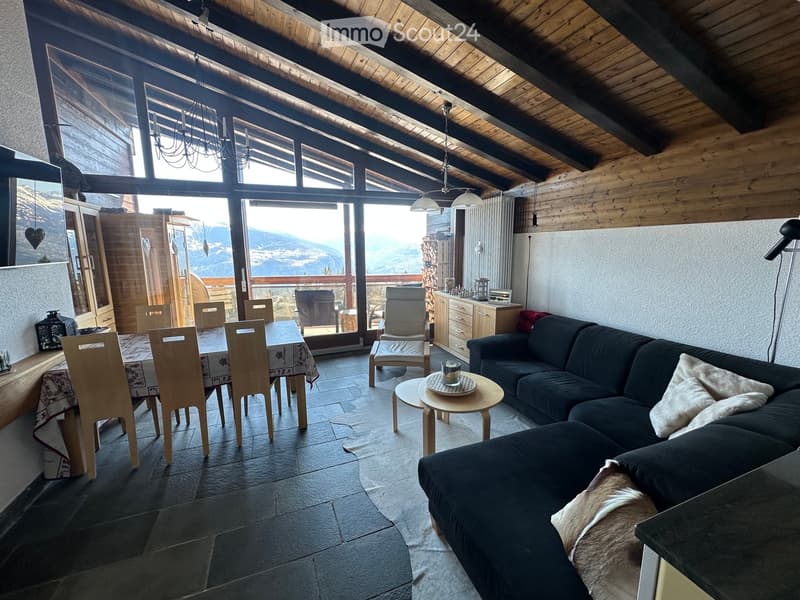 A spacious living and dining area with exposed wooden beams, large windows offering a scenic mountain view, a dark green sectional sofa, a small round coffee table, a dining table with chairs, and a cabinet with decorative items.