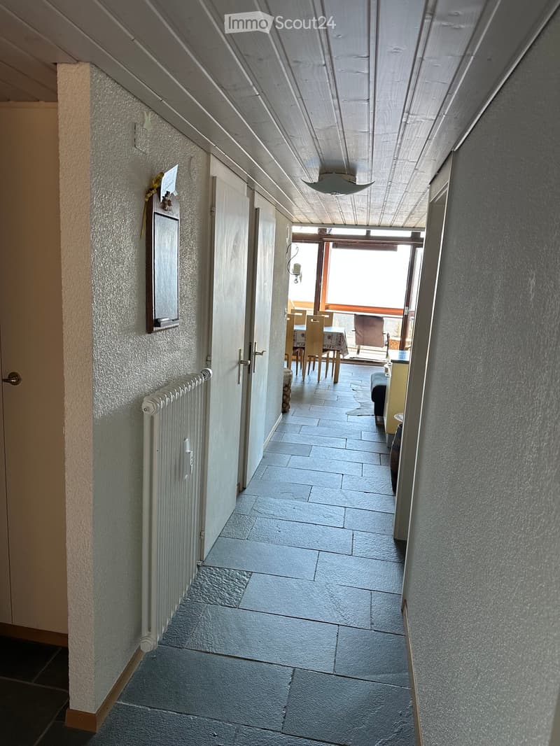 A narrow hallway with stone tiled flooring, white walls, and a radiator on the left side. The hallway leads to a room with a dining table and chairs visible at the end.