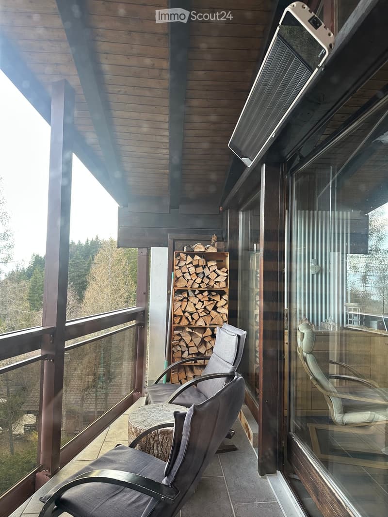 Wooden balcony with grey chairs, a round table, and a stack of firewood. Large glass windows and wooden railings.