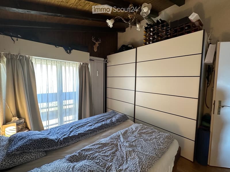 Cozy bedroom with a double bed, patterned bedding, large white wardrobe, window with curtains, radiator, nightstand with lamp, and a wine rack on top of the wardrobe.