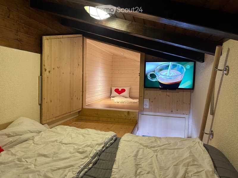 Cozy wooden bedroom with a large bed, a small seating area with a TV, and a heart-shaped pillow on a small bed.