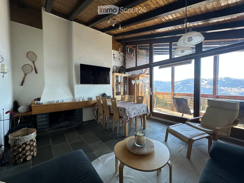 Bright living room with large windows offering a scenic mountain view, featuring a sofa, chairs, a coffee table, a dining area with a table and chairs, a fireplace, and a television mounted on the wall.