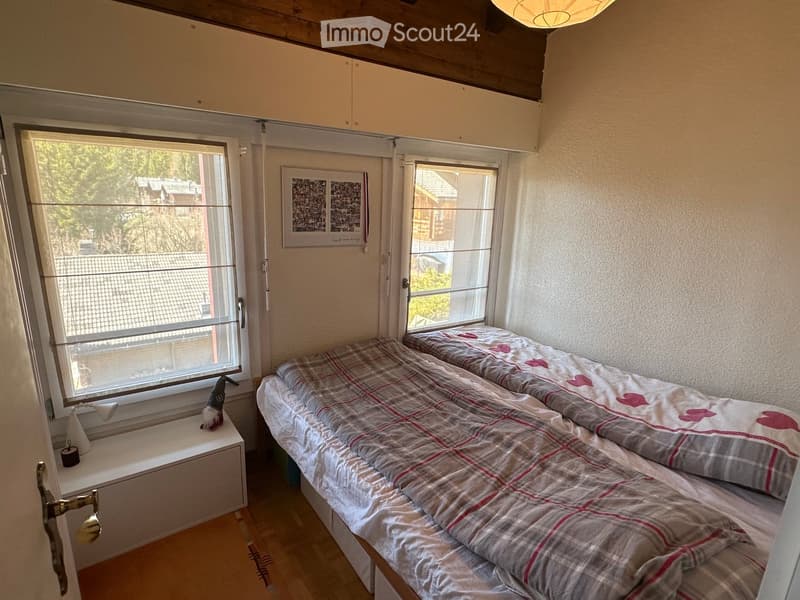 Cozy bedroom with two single beds, plaid bedding, large windows, and a small cabinet with a decorative item on top.