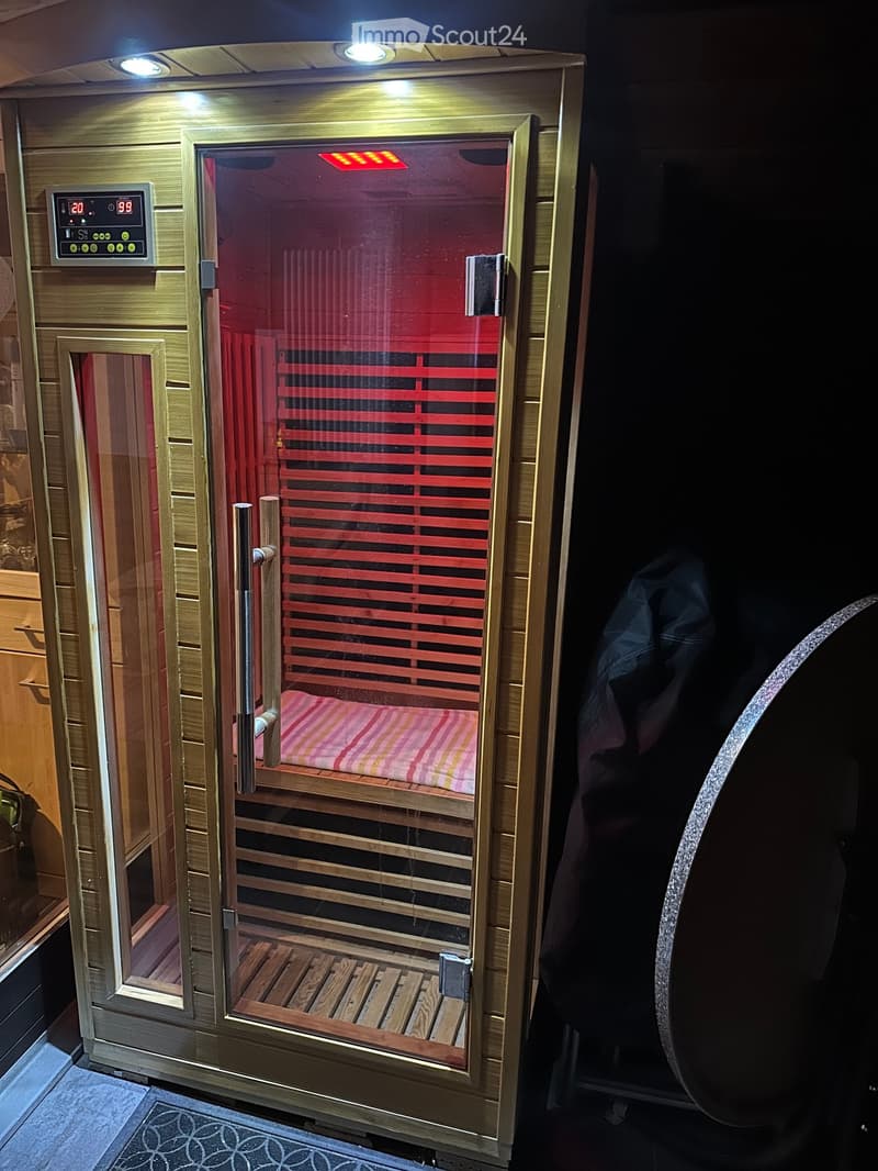 A modern sauna with wooden interior, glass door, control panel, and a striped cushion on the bench inside.