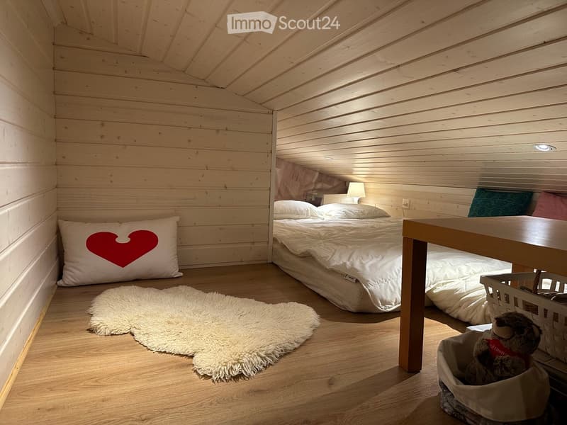 Cozy wooden bedroom with a double bed, nightstand, lamp, and a small table. Features a heart-shaped pillow and a plush toy in a basket.