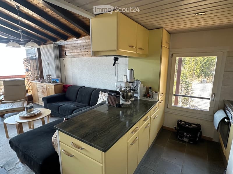 A modern, open-plan living and kitchen area with a dark grey sofa, a small round wooden table with a cup, a kitchen counter with cabinets, a mixer, and a refrigerator. The room has wooden beams on the ceiling and large windows offering a view of greenery.