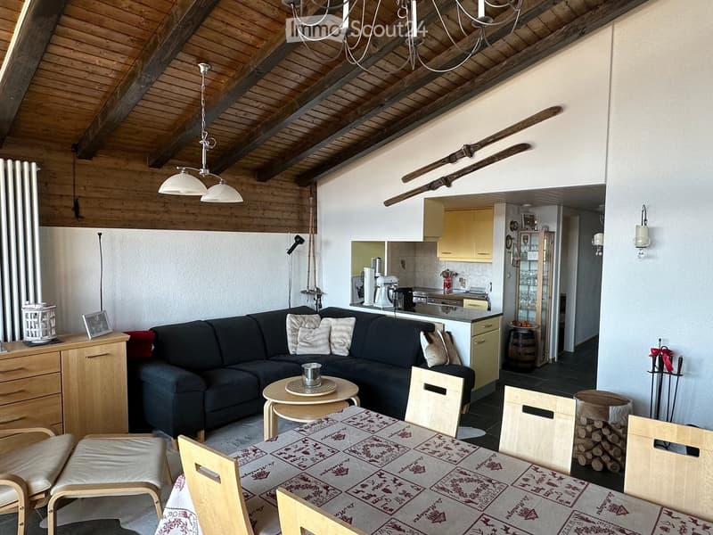 A cozy living room with a dark blue sectional sofa, wooden coffee table, dining table with chairs, and a small kitchen area in the background. The room features exposed wooden beams on the ceiling, pendant lights, and a radiator on the wall.
