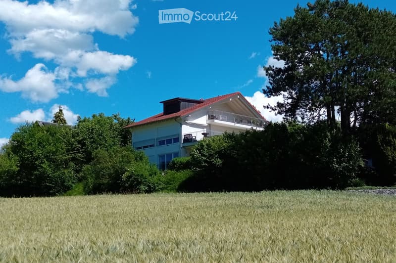 Two-story house with a red-tiled roof, white exterior, and a balcony on the upper floor, surrounded by trees and a grassy field.