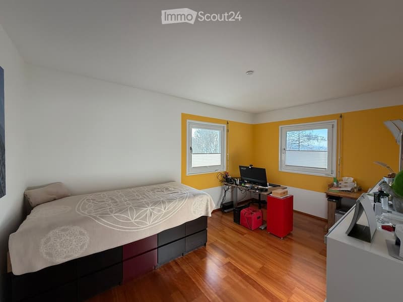Bright bedroom with wooden flooring, white and yellow walls, two windows, a bed with a patterned duvet, a desk with a laptop, and a small red stool.