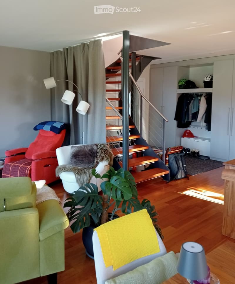 A modern living room with wooden flooring, a staircase leading to an upper level, and several pieces of furniture including a green sofa, a red armchair, a white chair with a fur throw, a small table with a yellow cushion, and a floor lamp. There is also
