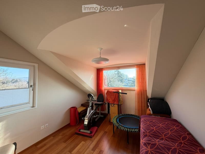 A cozy attic room with wooden flooring, large windows, a bed, exercise bike, and a small trampoline. The room features a slanted ceiling and warm lighting.