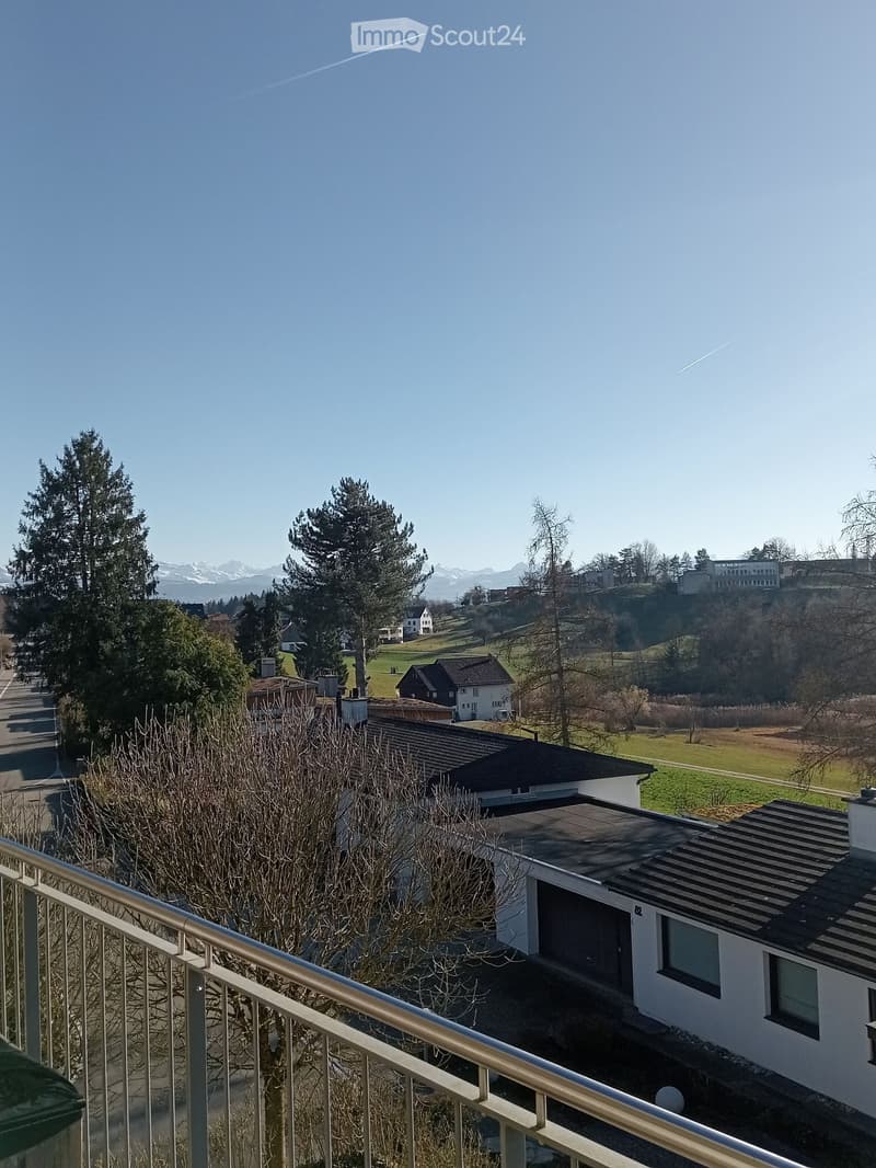 Balcony overlooking a scenic landscape with trees, houses, and distant mountains under a clear blue sky.