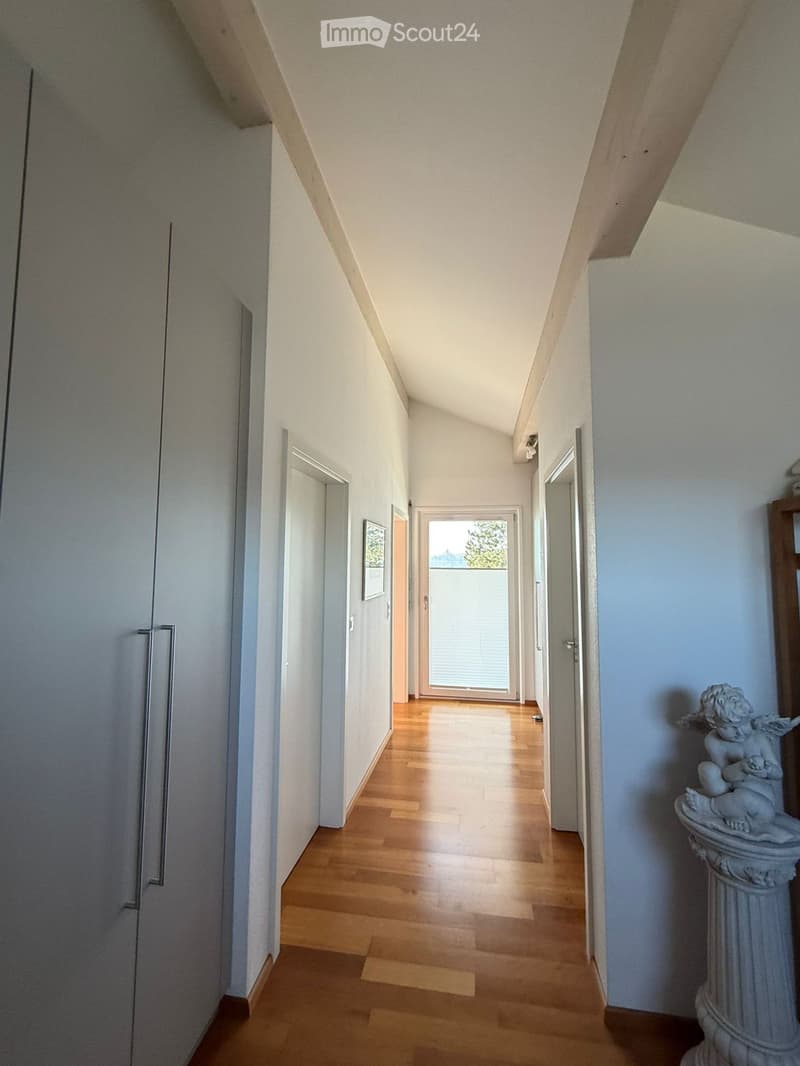 A long, narrow hallway with wooden flooring, white walls, and several doors leading to different rooms. Natural light streams in from a window at the end of the hallway.