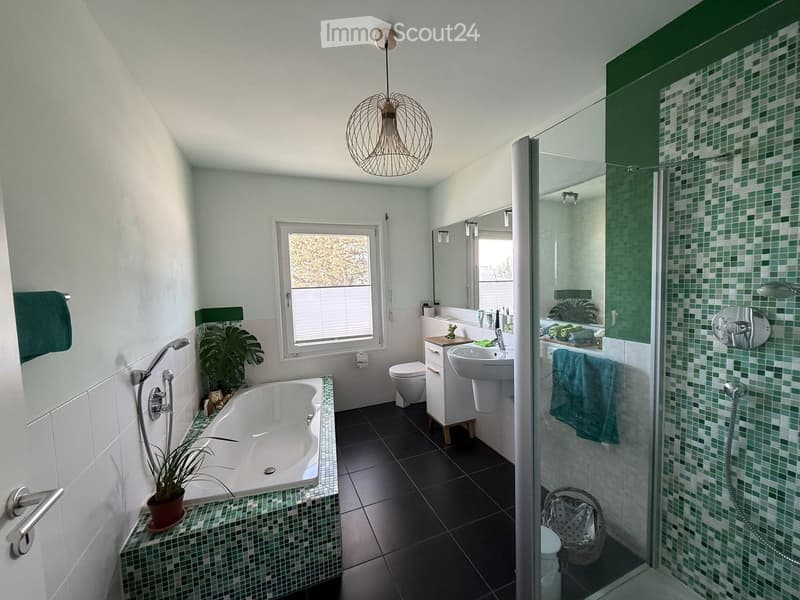 Modern bathroom with a bathtub, toilet, sink, shower, and decorative plant. Features green mosaic tiles and a hanging light fixture.