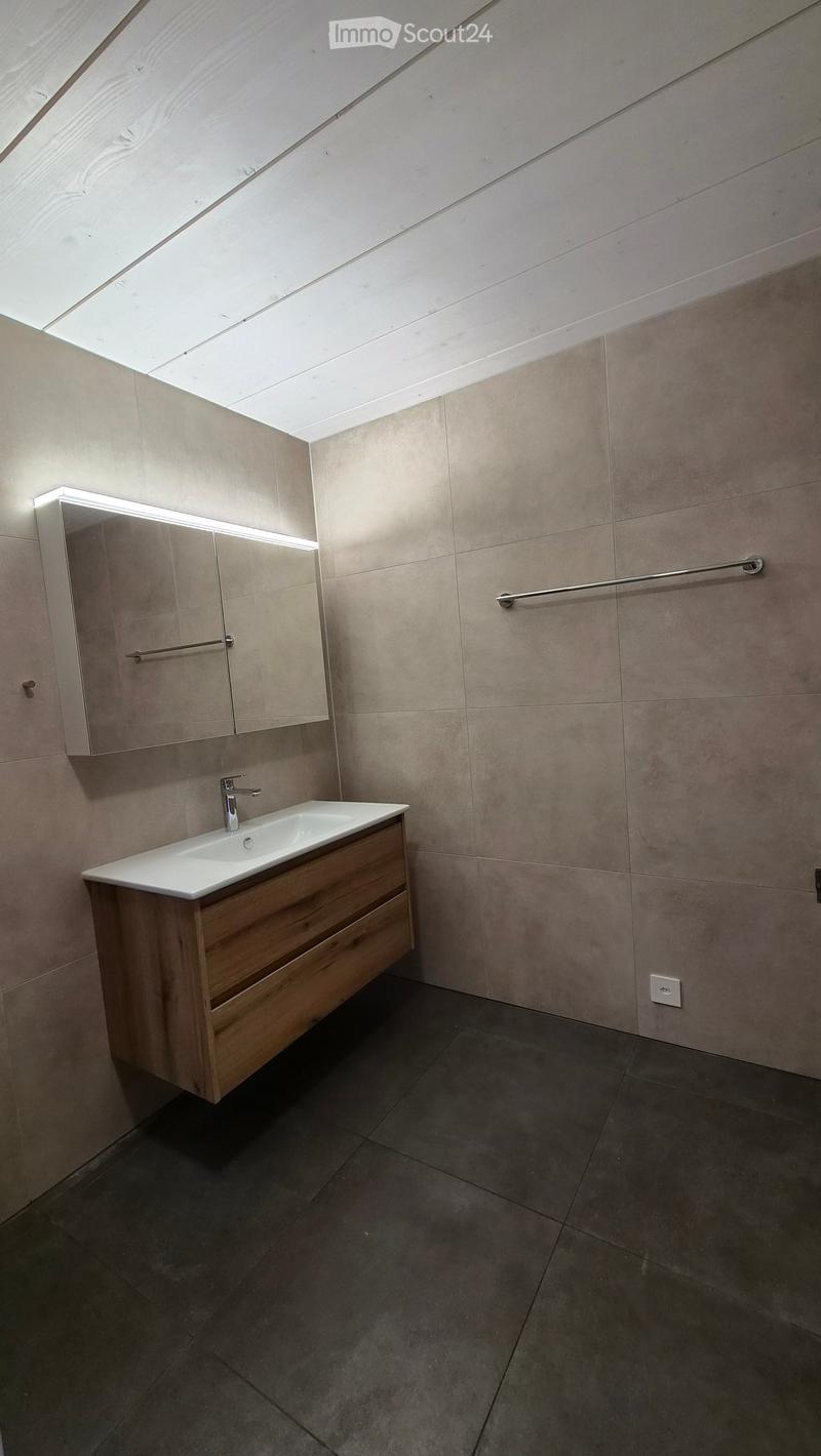 Modern bathroom with a white sink, wooden cabinet, mirror, and sleek faucet. The walls are tiled in a light beige color, and the floor is dark grey.