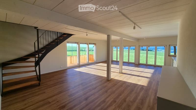 Open, empty space with wooden flooring, large windows, and a staircase leading to an upper level.