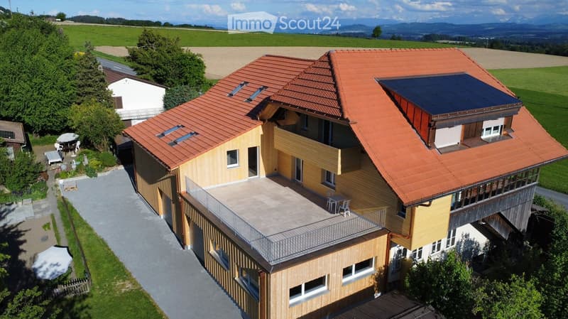 Modern two-story house with red-tiled roof, solar panels, balcony, and surrounding greenery.