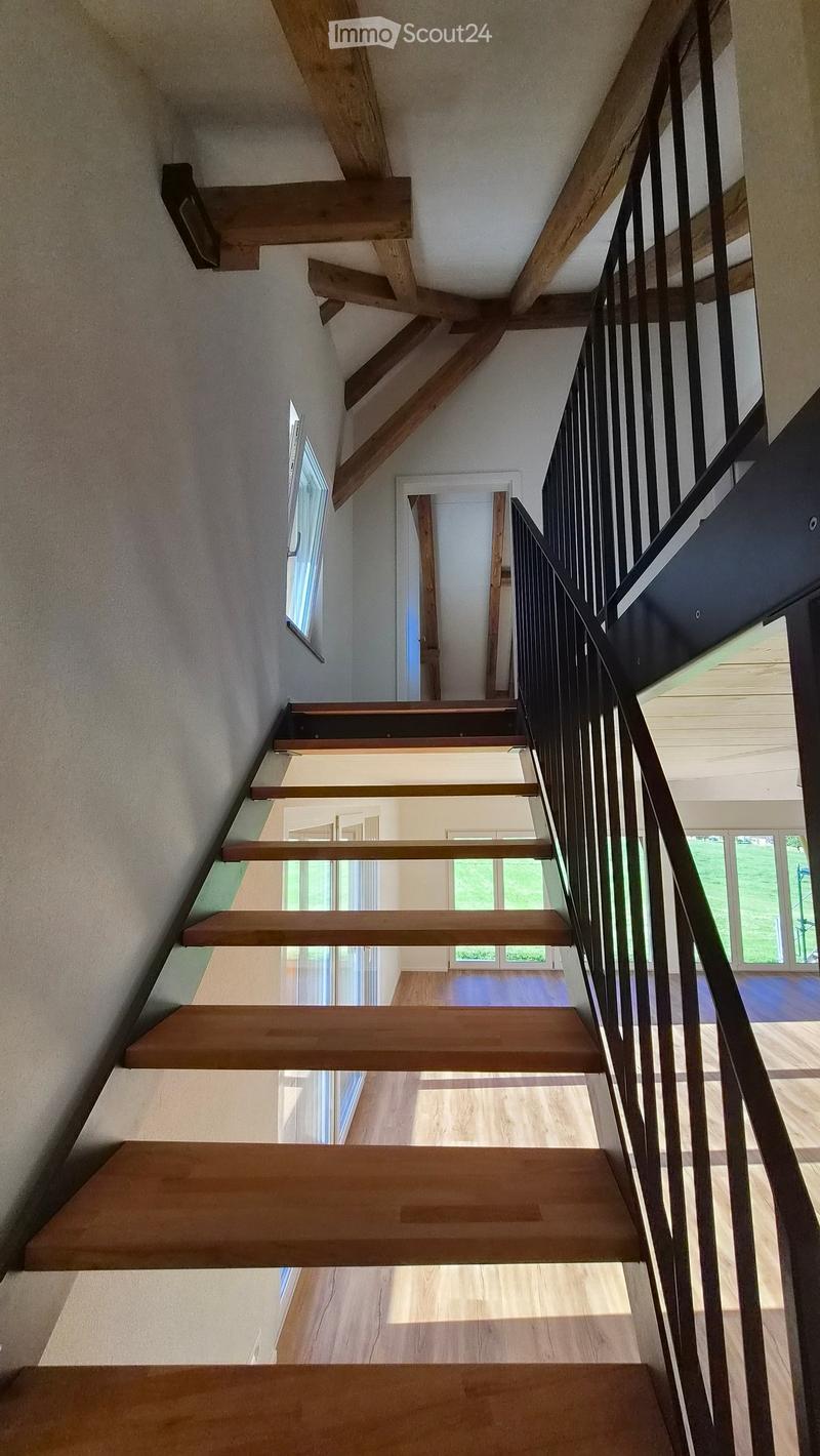 Wooden staircase with metal railing, light-colored walls, and a view of a room below with wooden flooring and large windows.