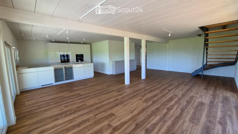 Open-plan space with light-colored wooden flooring, white cabinetry, and a staircase leading to an upper level. The area is unfurnished and appears to be a combined living and kitchen area.