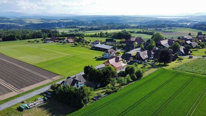 Aerial view of a rural village with scattered houses, green fields, and a road running through the area.