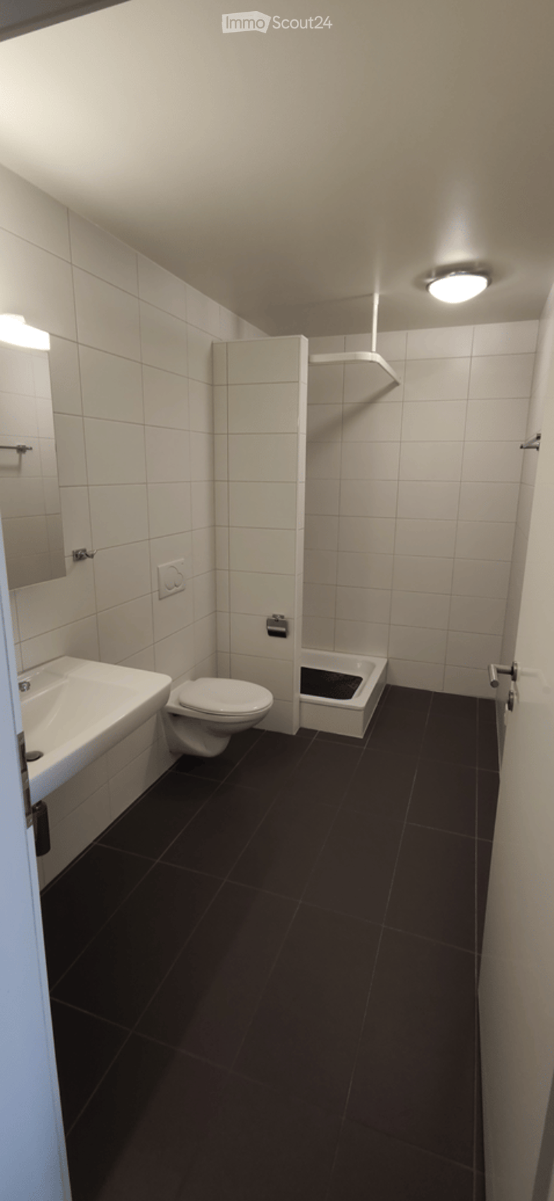 Modern bathroom with white tiled walls, black tiled floor, a sink, toilet, and a small shower stall. Features a mirror and a ceiling light.