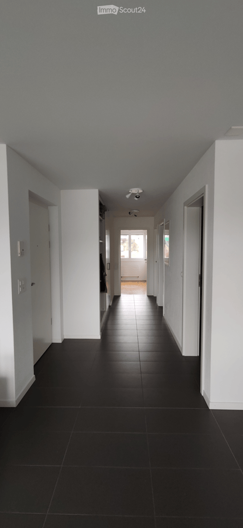 A long, narrow hallway with black tiled flooring and white walls. The ceiling is painted light blue, and there are several doors along both sides leading to other rooms. The hallway ends with a doorway that opens to an outdoor area.