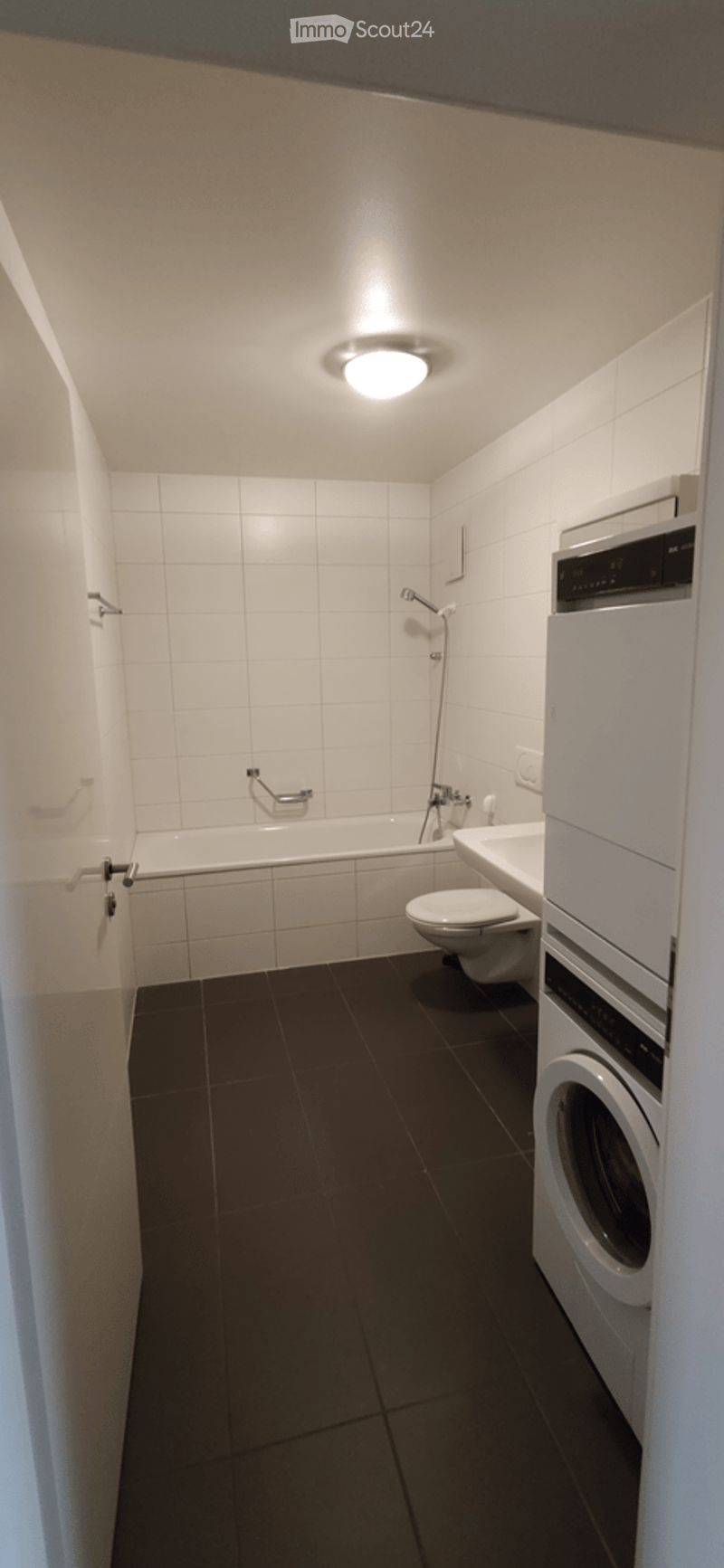 Modern bathroom with white tiled walls, dark tiled floor, bathtub, toilet, sink, shower, and a private washing machine.