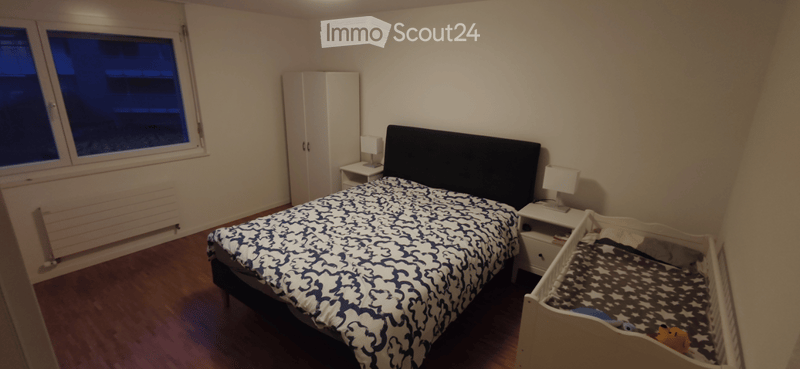Cozy bedroom with a double bed, nightstands, a wardrobe, and a small crib. The room features wooden flooring and large windows.
