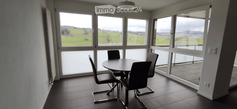 Bright dining area with large windows offering a scenic view of green fields and hills. The room features a round table with four black chairs, tiled flooring, and modern lighting.