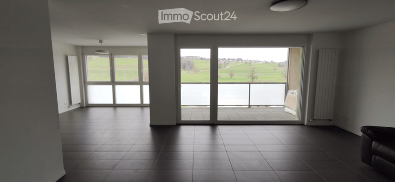 Empty room with large windows and a sliding glass door leading to a balcony, overlooking a green field.