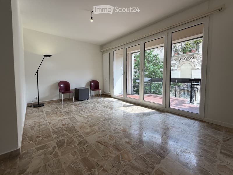 Bright, unfurnished room with large sliding glass doors leading to a balcony, stone tile flooring, and minimal furniture including two chairs, a small table, and a floor lamp.