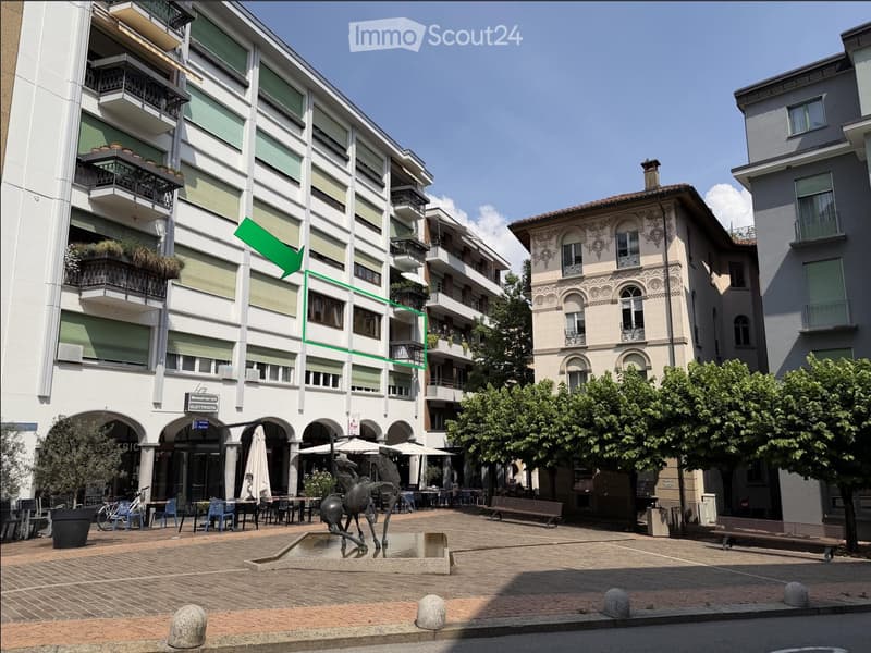 Multi-story building with balconies and green shutters, located in an urban area with outdoor seating, a fountain, and trees.