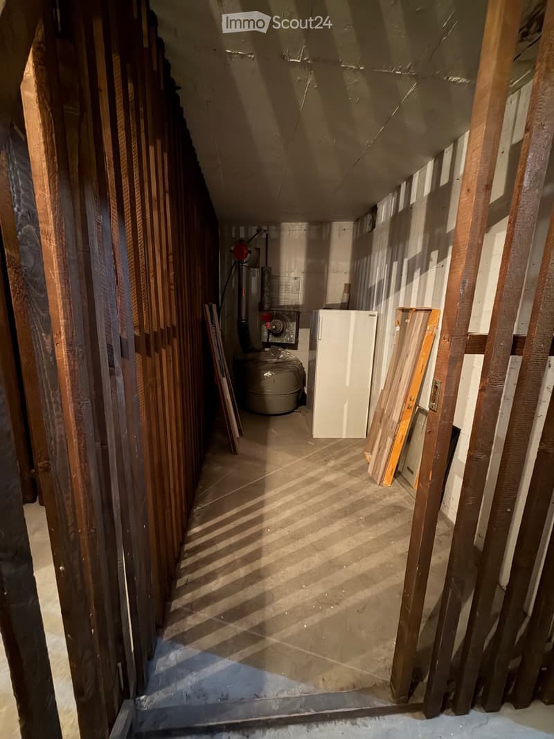 Empty basement space with exposed wooden beams, concrete floor, and minimal furnishings including a water heater, refrigerator, and some stacked materials.