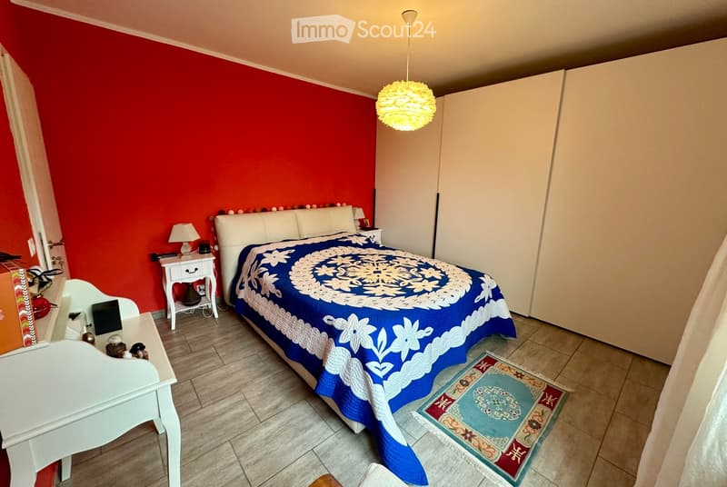 Bedroom with red accent wall, white bed with blue and white patterned bedspread, nightstand with lamp, small table with items, and a decorative rug on the floor.
