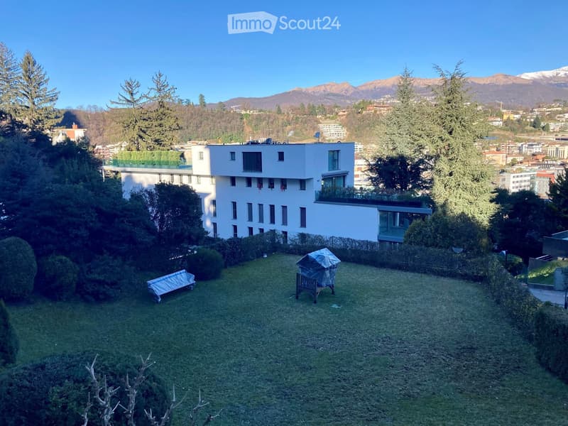 A well-maintained garden area with a modern white building in the background, surrounded by trees and a scenic mountain view.