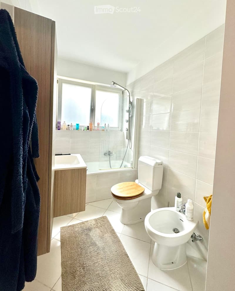 Modern bathroom with white tiles, a toilet with a wooden seat, a sink, a bathtub with a glass shower enclosure, a wooden cabinet, a bathrobe, and a towel.