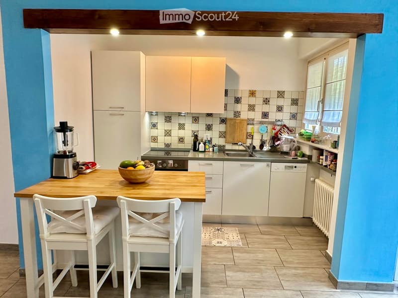 Bright kitchen with white cabinets, wooden island, two white chairs, bowl of fruit, blender, sink, and dishwasher.
