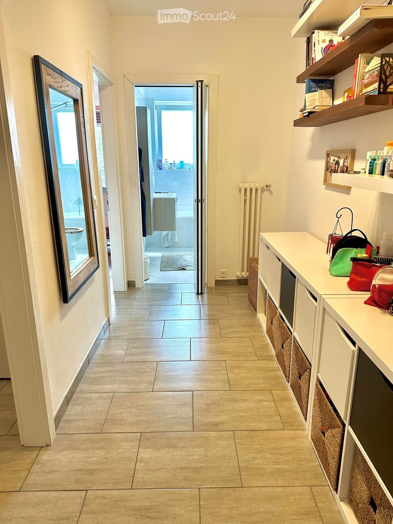 A clean, tiled hallway with a white cabinet on the right side, a mirror on the left, and an open doorway leading to a bathroom with a toilet and bathtub visible.
