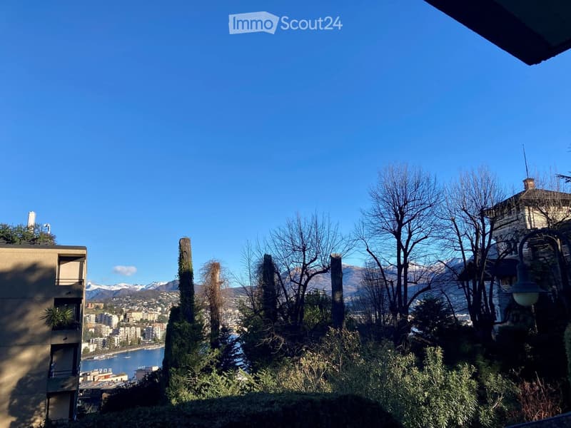 A scenic outdoor view from a balcony or terrace, showing a cityscape with buildings, a body of water, and distant mountains under a clear blue sky.