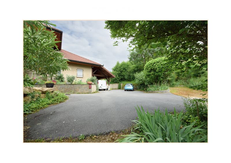 A two-story house with a red-tiled roof, surrounded by lush greenery and a driveway leading to a covered entrance area. A white van and a blue car are parked near the house.