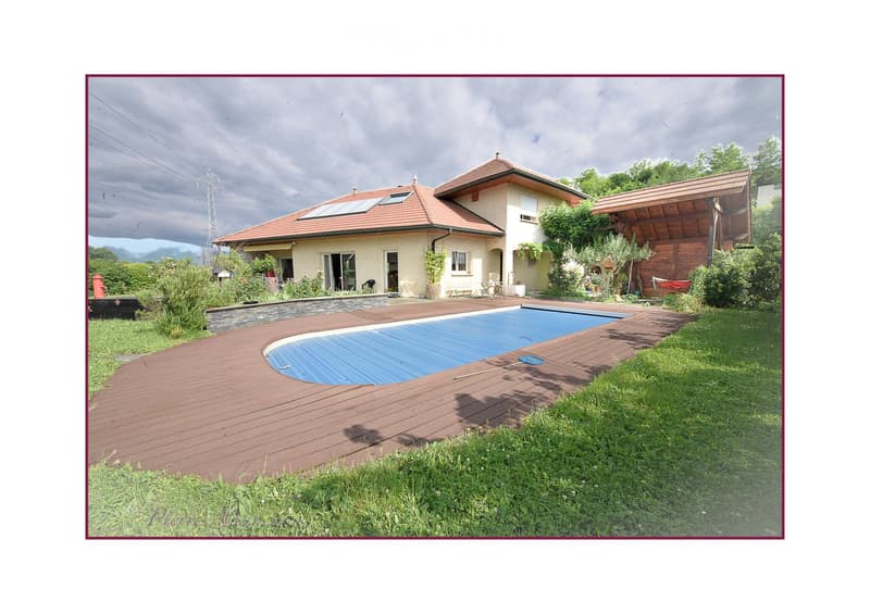 Outdoor area with a swimming pool, wooden decking, and surrounding greenery, featuring a house with a tiled roof and solar panels in the background.