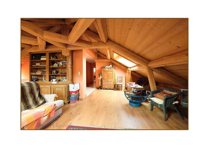 Cozy attic room with wooden beams, exposed wooden ceiling, and warm lighting. Features a sofa, armchair, small table, bookshelf, and a mirror.