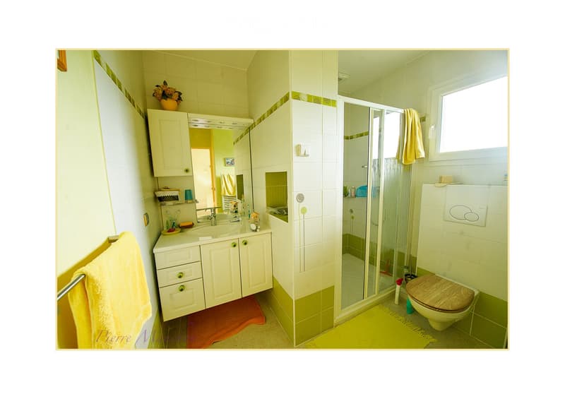 Bright bathroom with white tiles, a sink with a mirror cabinet, a shower area, and a toilet with a wooden seat. Towels and a flower vase are visible.